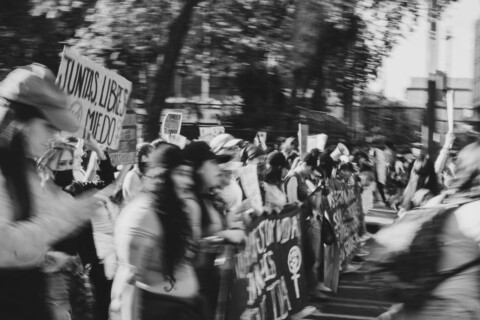 Segmento di manifestazione femminista, persone tengono in mano uno striscione, foto in bianco e nero con effetto mosso che rende poco chiari i volti