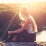 Una ragazza bionda seduta di spalle guarda un fiume, il sole le illumina i capelli