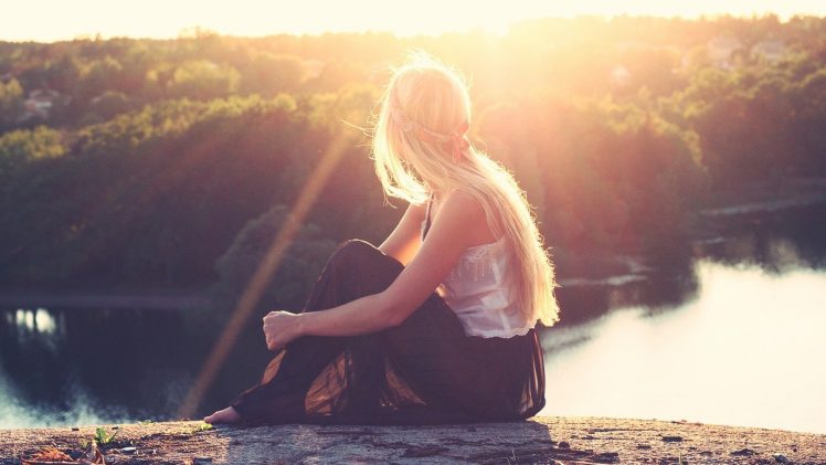 Una ragazza bionda seduta di spalle guarda un fiume, il sole le illumina i capelli