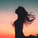 Profilo di una donna in controluce, con i capelli lunghi che fluttuano nel vento. Sullo sfondo, un'alba rosseggiante